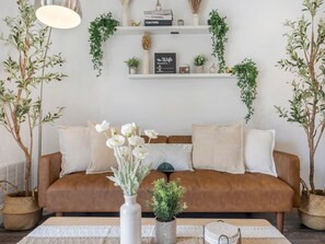Una sala de estar con un sofá de cuero marrón, cojines blancos, un jarrón blanco con flores blancas y una pequeña planta en maceta sobre una mesa de madera.