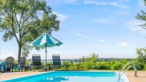 A swimming pool with lounge chairs and an umbrella, surrounded by trees and a clear sky.