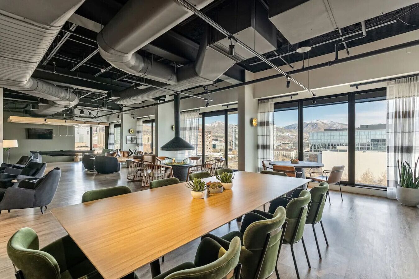 A modern office space with a large wooden conference table, green upholstered chairs, and large windows offering a view of a cityscape and mountains.