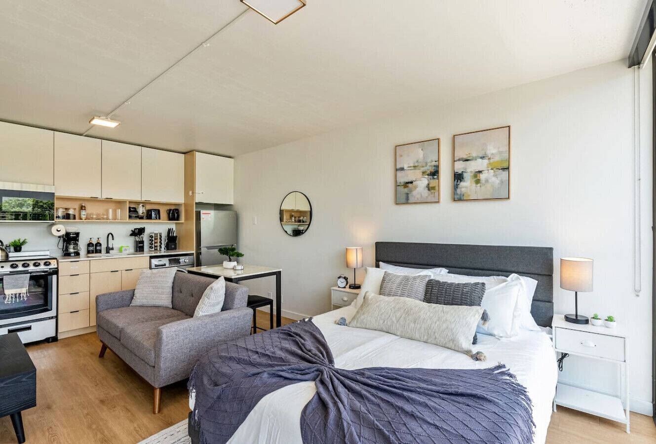 A modern bedroom with a large bed, a sofa, a desk, and a kitchen area.