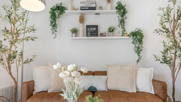 Una sala de estar con un sofá de cuero marrón, cojines blancos, un jarrón blanco con flores blancas y una pequeña planta en maceta sobre una mesa de madera.