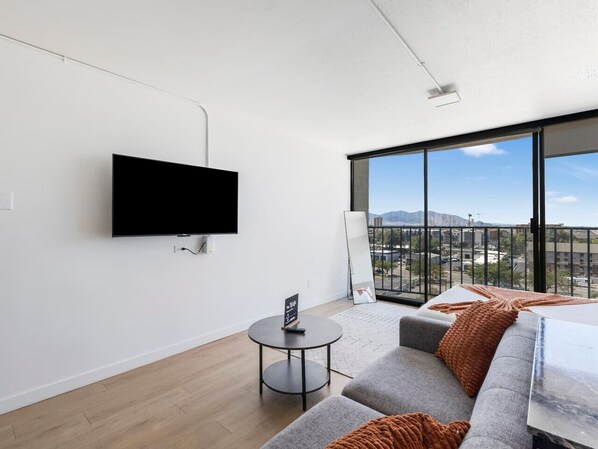 A modern hotel room with a large window, a flat-screen TV mounted on the wall, a gray sofa, a small round table, and a view of a cityscape.