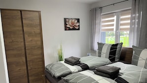 Een slaapkamer met een bed, kussens, een houten garderobe, een raam met gordijnen en een aan de muur hangend schilderij.