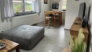 Een moderne woonkamer met een grijze bank, een houten eettafel met stoelen, een televisie en een keukenhoek met een fornuis en magnetron.