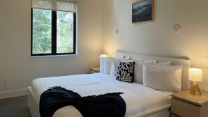 A bedroom with a bed, bedside tables, a nightstand, a window, and a wall-mounted picture. - Cliffside Hideaway: Luxurious, Peaceful, with Gorgeous views Over the Ocean (Port Renfrew)