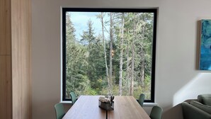A modern dining area with a wooden table, green chairs, and a large window overlooking a forest. - Cliffside Hideaway: Luxurious, Peaceful, with Gorgeous views Over the Ocean (Port Renfrew)