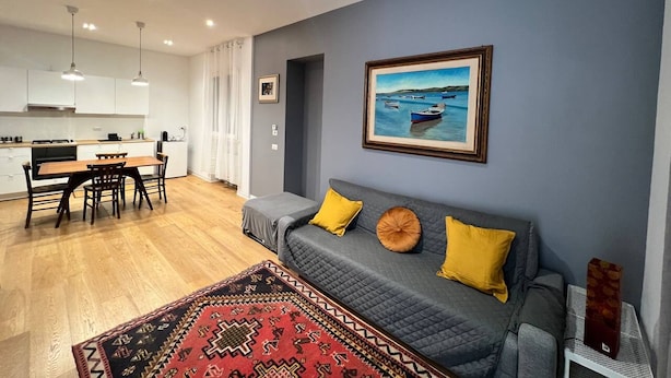 A modern living room with a grey sofa, yellow cushions, a patterned rug, and a painting of boats on the wall. - Pisu Apartments (appart.Orsolina)  (Quartucciu)