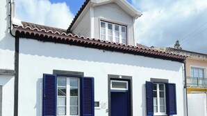 A two-story building with a white facade and a tiled roof. There are two windows with blue shutters, one of which has a door.