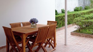 A covered patio with a wooden table and chairs, a vase with flowers, and a view of a garden and building in the background.