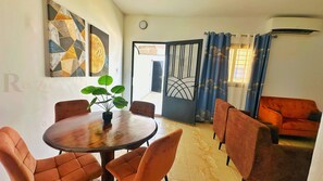 A dining area with a round table, six chairs, and a potted plant. There is a sofa and a window with curtains in the background. - Res'Zac (Rufisque)