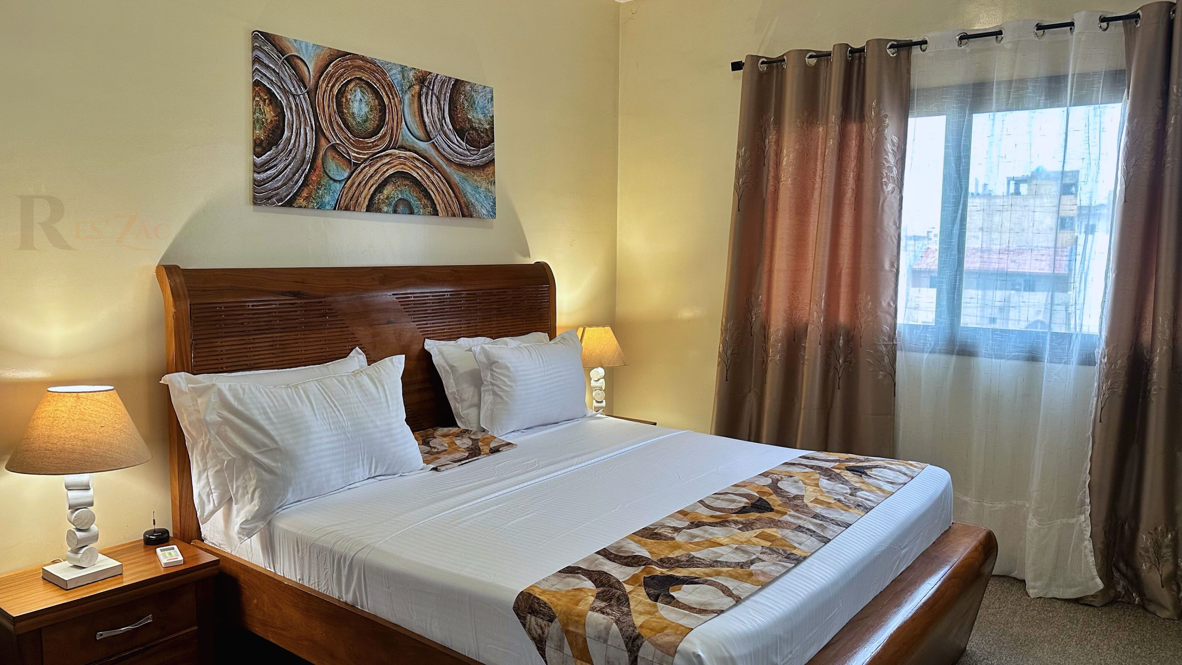 A neatly made bed with white linens and a patterned bedspread, a wooden headboard, a bedside table with a lamp, and a wall-mounted artwork.