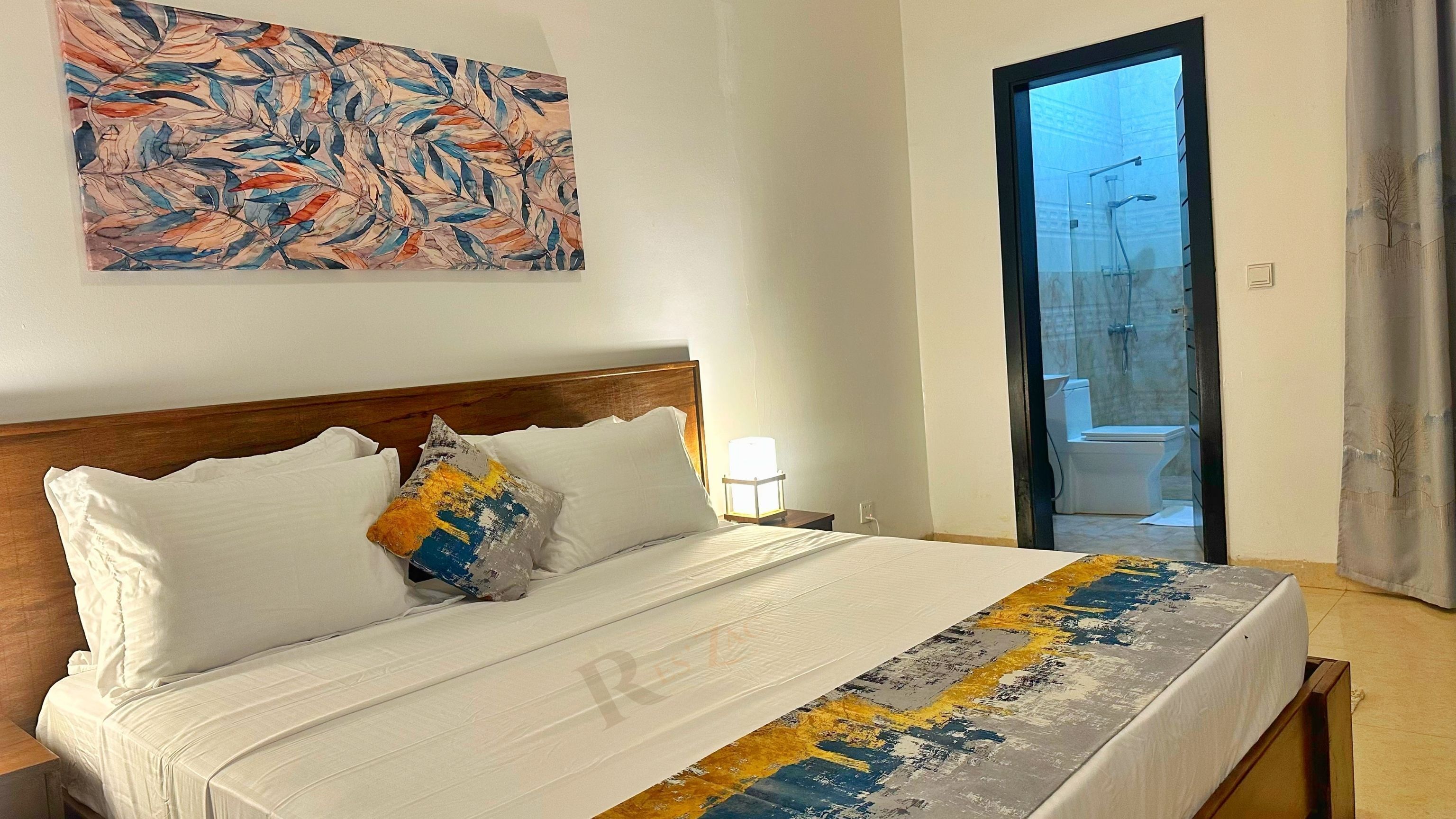 Une chambre d’hôtel avec un lit, une table de chevet, une lampe, une salle de bain visible à travers une porte ouverte, et un grand tableau abstrait accroché au mur.