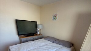 Ein Schlafzimmer mit einem Bett, einem Fernseher auf einem Holztischchen, einer Wanduhr und einer hängenden Glühbirne.