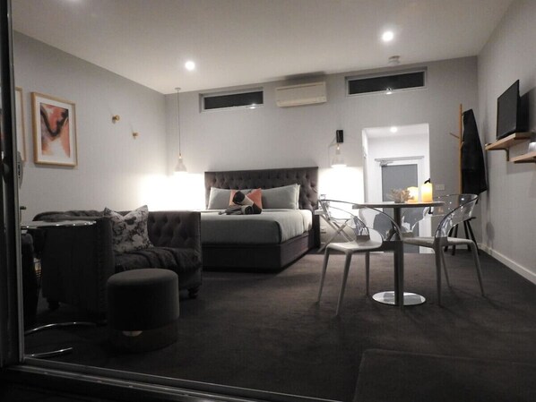 A modern bedroom with a large bed, a sofa, a glass dining table, and a TV.