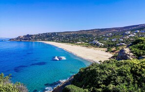 Ein Küstenblick mit Sandstrand, klarem blauen Wasser und einer Hügelslandschaft mit Gebäuden.