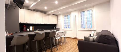 A modern kitchen with a bar area, wooden flooring, and a sofa in a living area with windows.