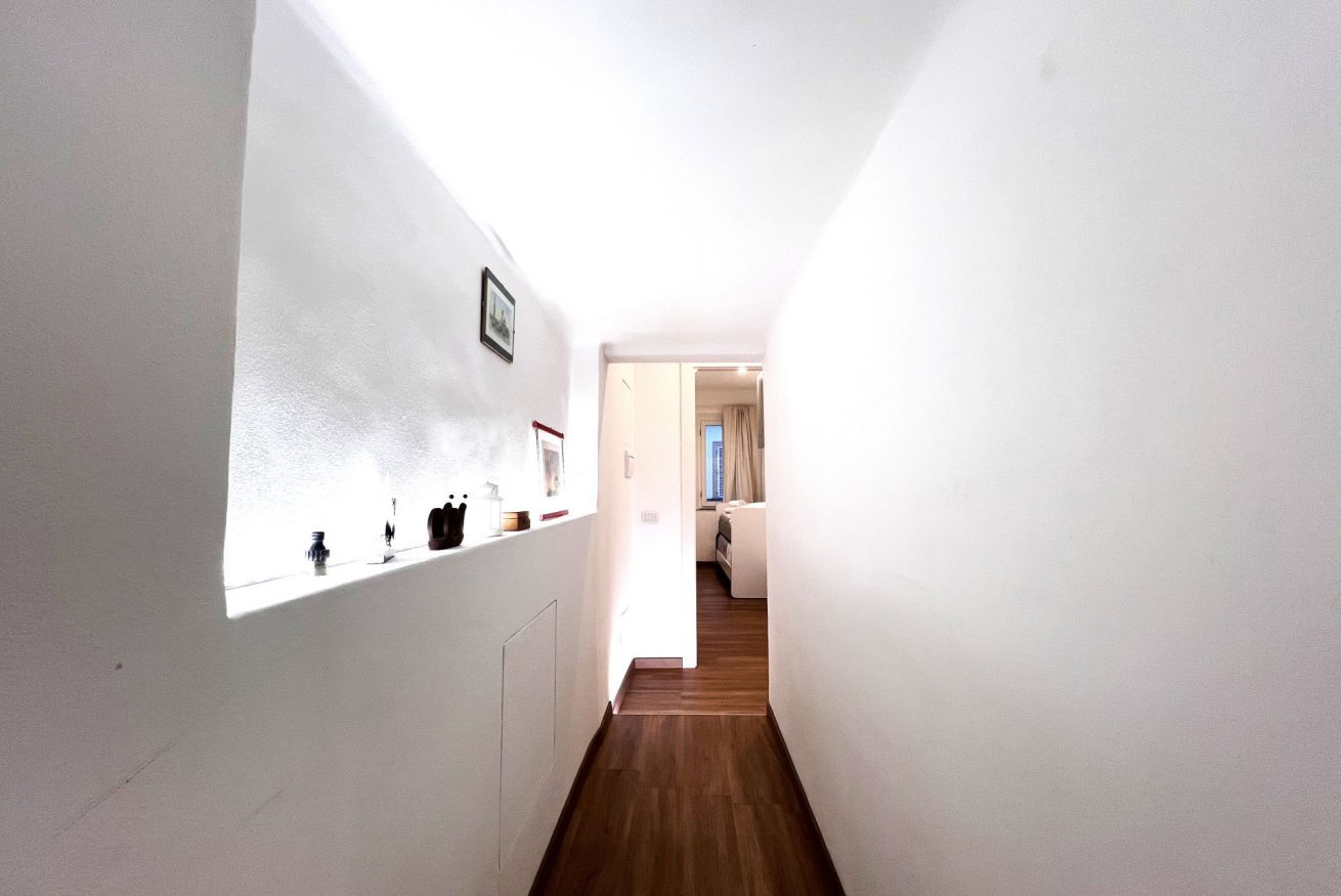 Un corridoio stretto con pareti bianche e pavimento in legno che conduce a una camera da letto con un letto e una finestra.