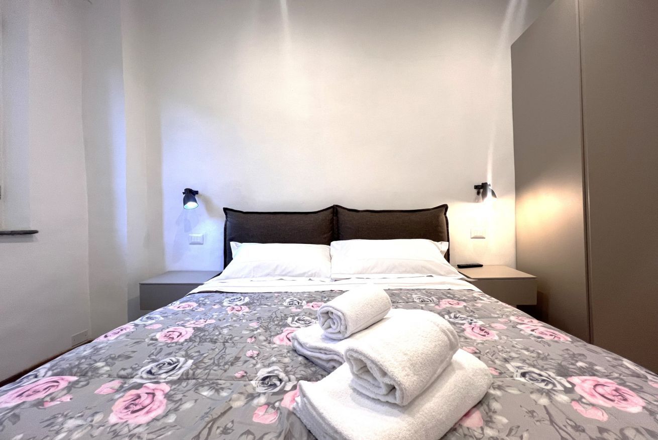 A neatly made bed with a floral bedspread, two bedside lamps, and rolled towels on the bed.