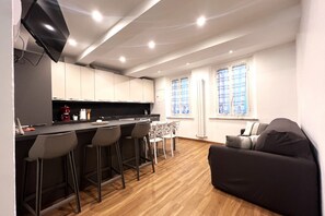 A modern kitchen with a bar area, wooden flooring, and a sofa in a living area with windows.