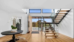 A modern interior with a wooden floor, a glass staircase, a round table with a plant, and a glass door leading outside.