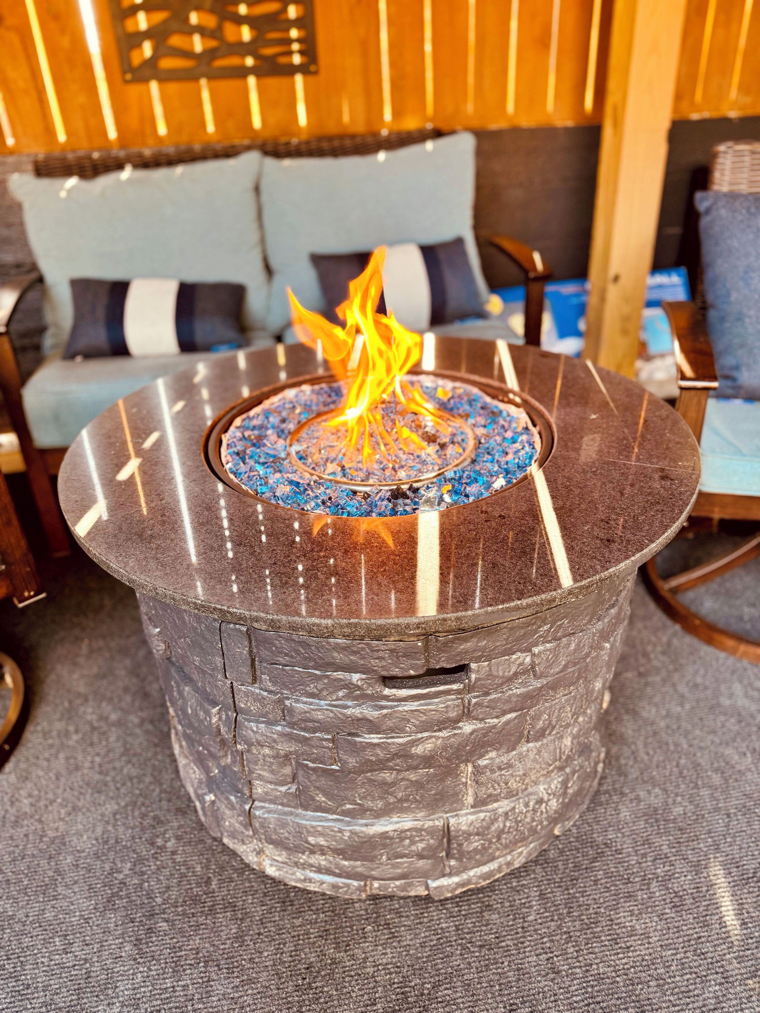 A stone fire pit with a blue glass top, surrounded by a grey textured floor, in a room with wooden walls and a sofa with striped cushions.