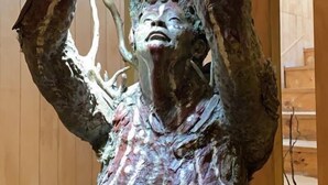 Une sculpture représentant une figure aux bras levés, placée sur un fond en bois avec un escalier.