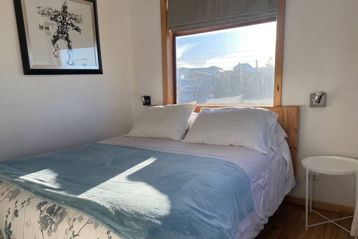 A bedroom with a bed, a window, a bedside table, and a framed picture on the wall.
