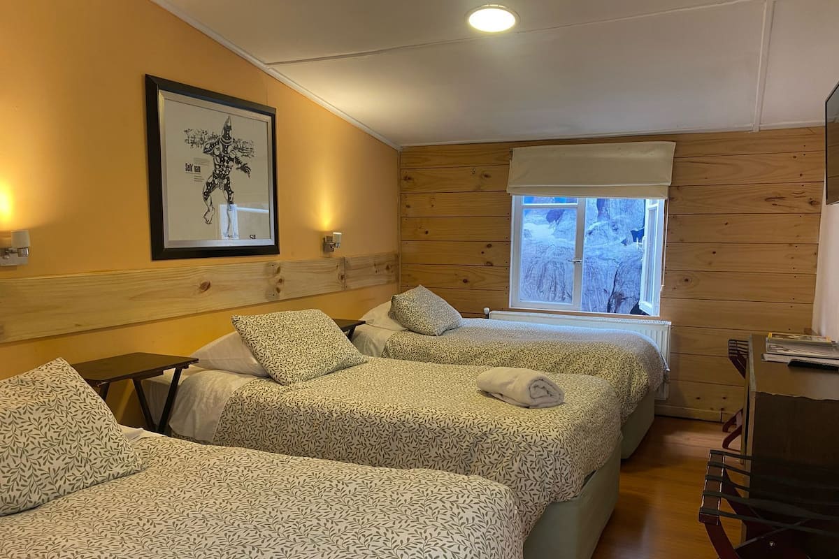 A room with two beds, a window with a view of a snowy landscape, a framed picture on the wall, and a small table with a chair.
