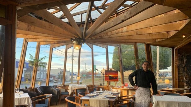 Un restaurant avec des poutres en bois et de grandes fenêtres donnant sur l’extérieur. Les tables sont dressées avec des assiettes et des verres, et une personne se tient près des fenêtres.