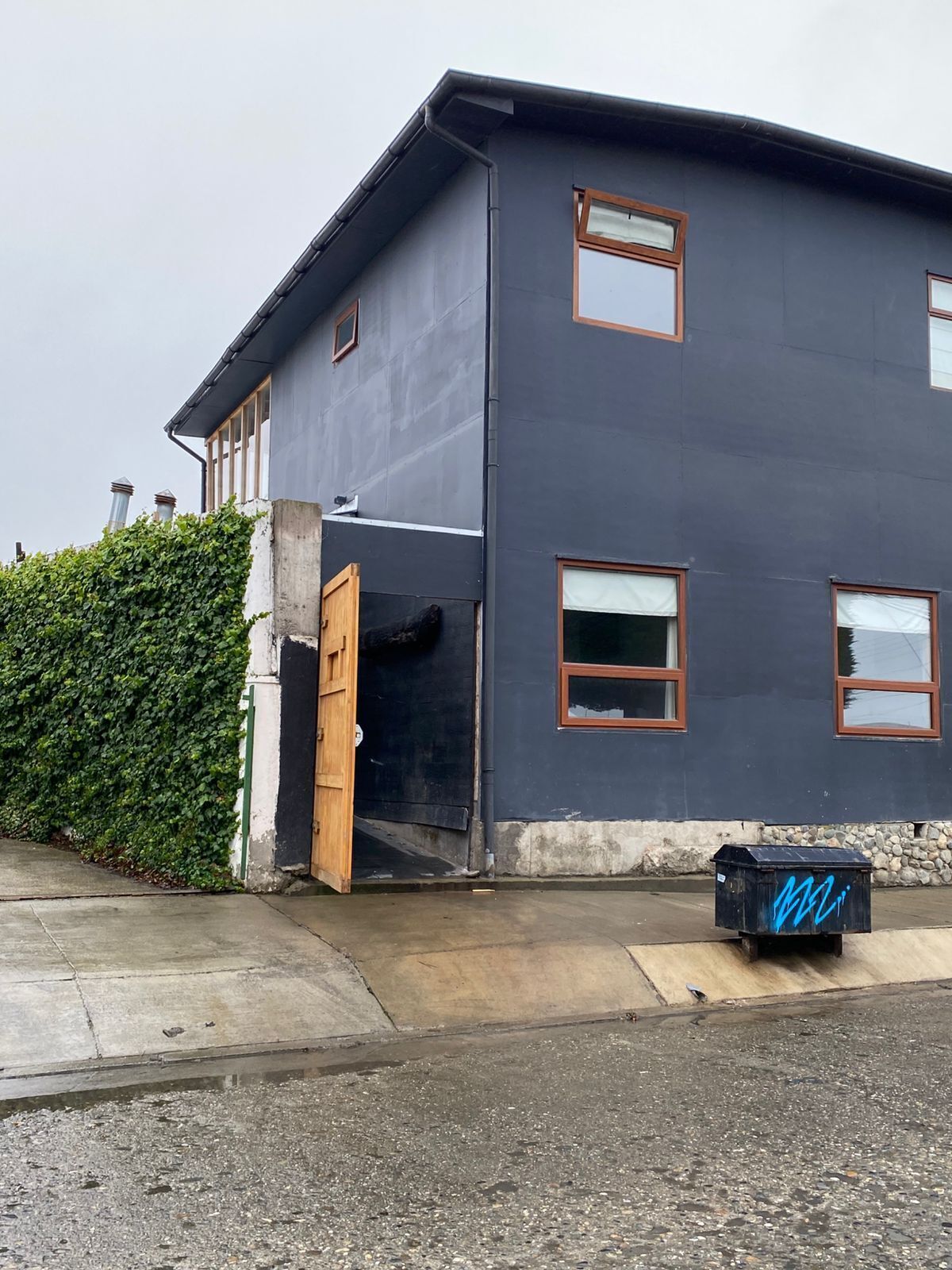 Un bâtiment à deux étages, doté d’une façade sombre, d’une porte en bois et d’une petite poubelle noire ornée de graffitis bleus.