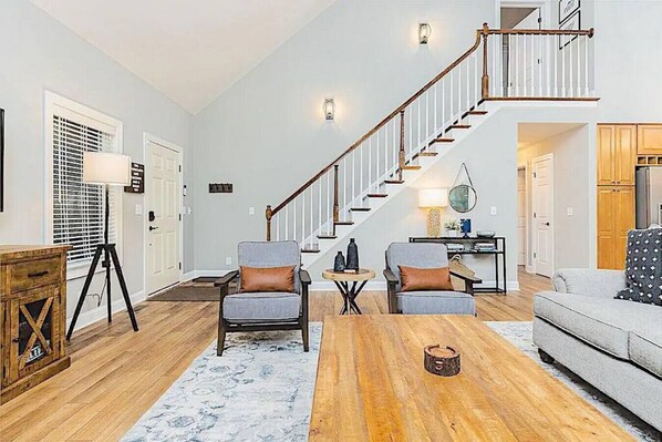 A spacious living room with a wooden floor, two armchairs, a sofa, a coffee table, and a staircase with a railing.