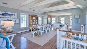 Een ruime eetruimte met een hoge plafond en zichtbare balken, een houten eettafel met witte stoelen en een groot raam dat natuurlijk licht binnenlaat.