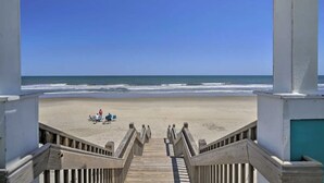 Een houten trap die naar een strand leidt, met een helder uitzicht over de oceaan en enkele mensen in de verte.