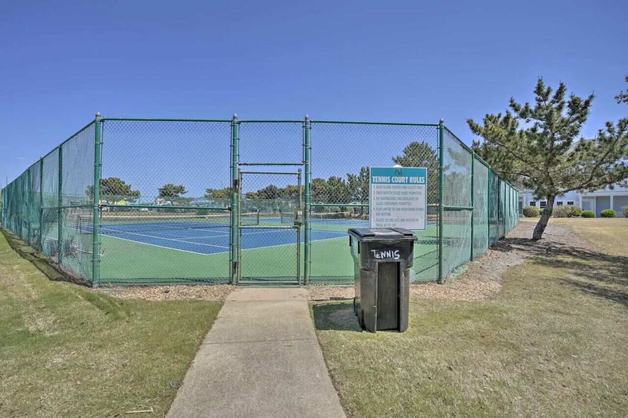 A tennis court enclosed by a green fence with a trash bin labeled "TENNIS" in front.