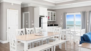 Un vaste espace de salle à manger avec une table en bois et des chaises blanches, une cuisine avec des meubles blancs et une vue sur l’océan grâce à de grandes fenêtres.