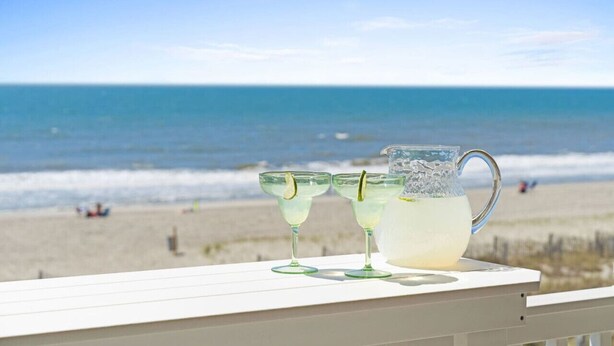 Une vue sur la plage avec une balustrade blanche, deux verres et une carafe de boisson posée sur la balustrade.