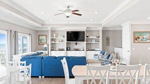 Un salon spacieux avec un canapé bleu, des chaises blanches, une table à manger et un ventilateur de plafond.