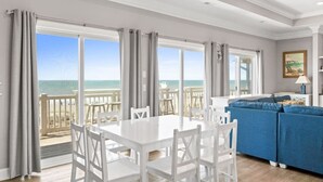 Une pièce avec une table et des chaises blanches, un canapé bleu et de grandes fenêtres donnant sur la plage.