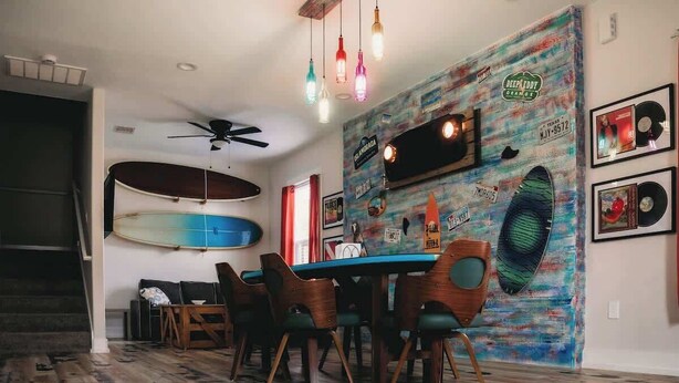 A dining area with a wooden table and chairs, a ceiling fan, and a wall decorated with surfboards and music memorabilia. - The Board House "Longboard" (Galveston)