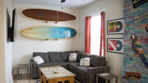 A living room with a sectional sofa, a coffee table, and two surfboards mounted on the wall. - The Board House "Longboard" (Galveston)