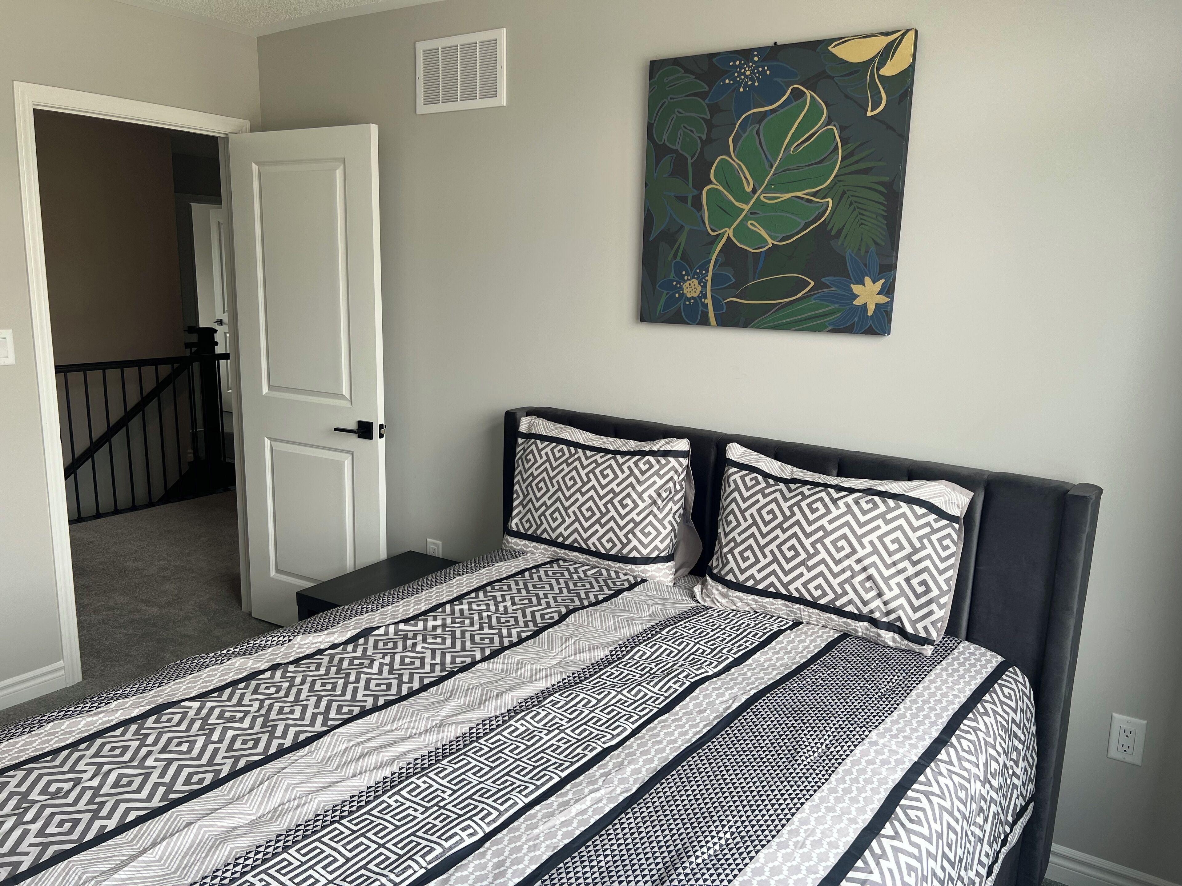 A bedroom with a bed, patterned bedding, a door, a staircase, and a wall with a floral painting.