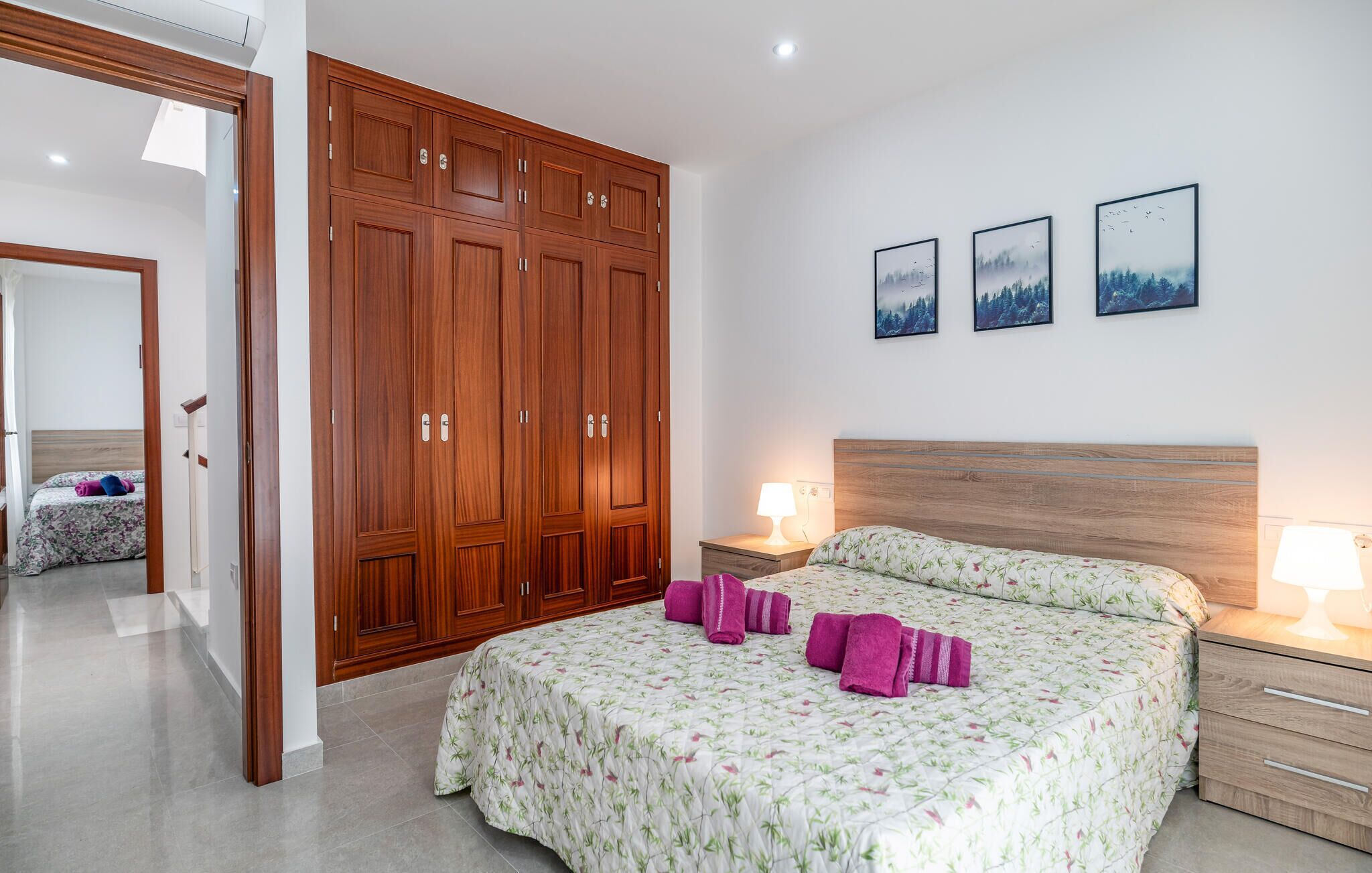 A bedroom with a large wooden wardrobe, a bed with floral bedding, two bedside lamps, and three framed pictures on the wall.