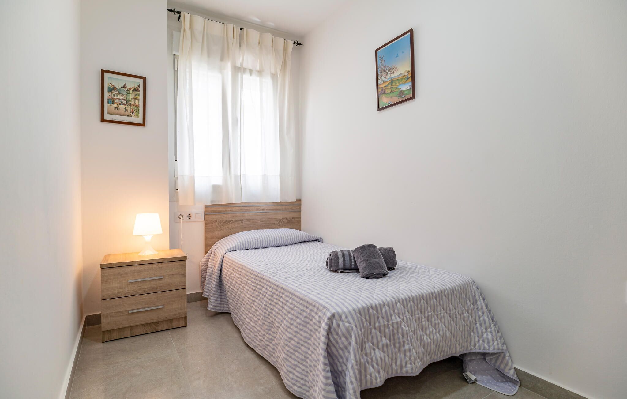 A bedroom with a bed, a nightstand with a lamp, a window with curtains, and two framed pictures on the wall.