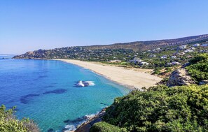 A coastal landscape with a clear blue sea, sandy beach, and a hillside with scattered buildings.