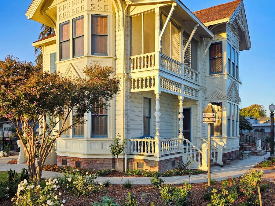 A two-story Victorian-style house with a wrap-around porch, a front entrance with a staircase, and a small front yard with flowers and shrubs.