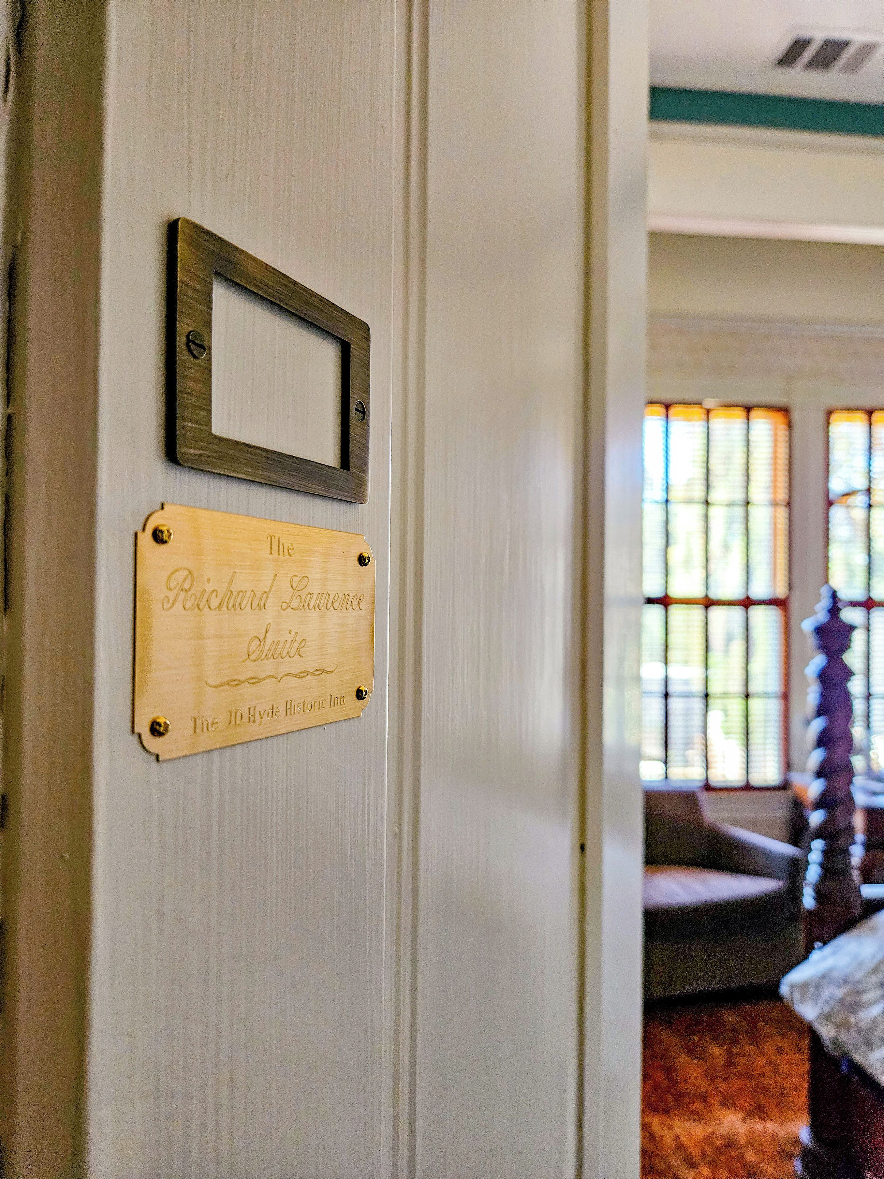 Una placa de madera con texto grabado en una pared, un pequeño espejo rectangular encima y una ventana con una silla en una habitación a medio ver.