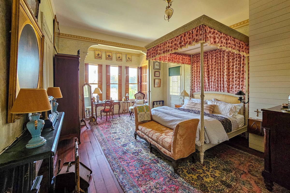 A traditional bedroom with a four-poster bed, a sitting area with a sofa and chairs, and a large mirror.