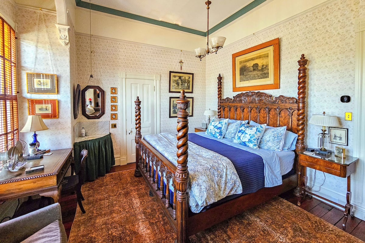 A four-poster bed with a floral bedspread, a wooden nightstand with a lamp, and a wall-mounted picture frame.