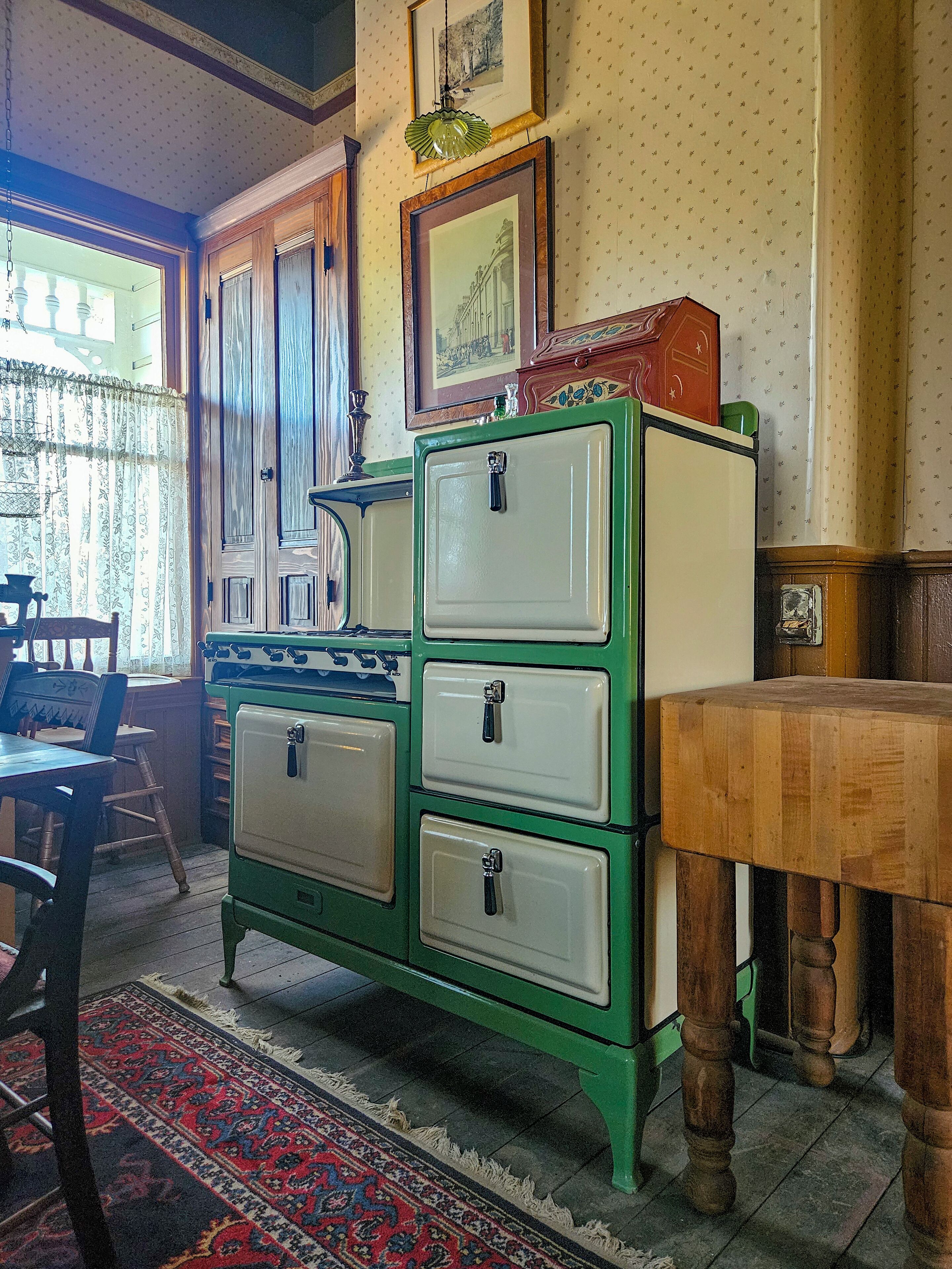 Een retro groene en witte keuken met een houten tafel en stoelen, een vloerkleed met dessin en een raam met gordijnen.