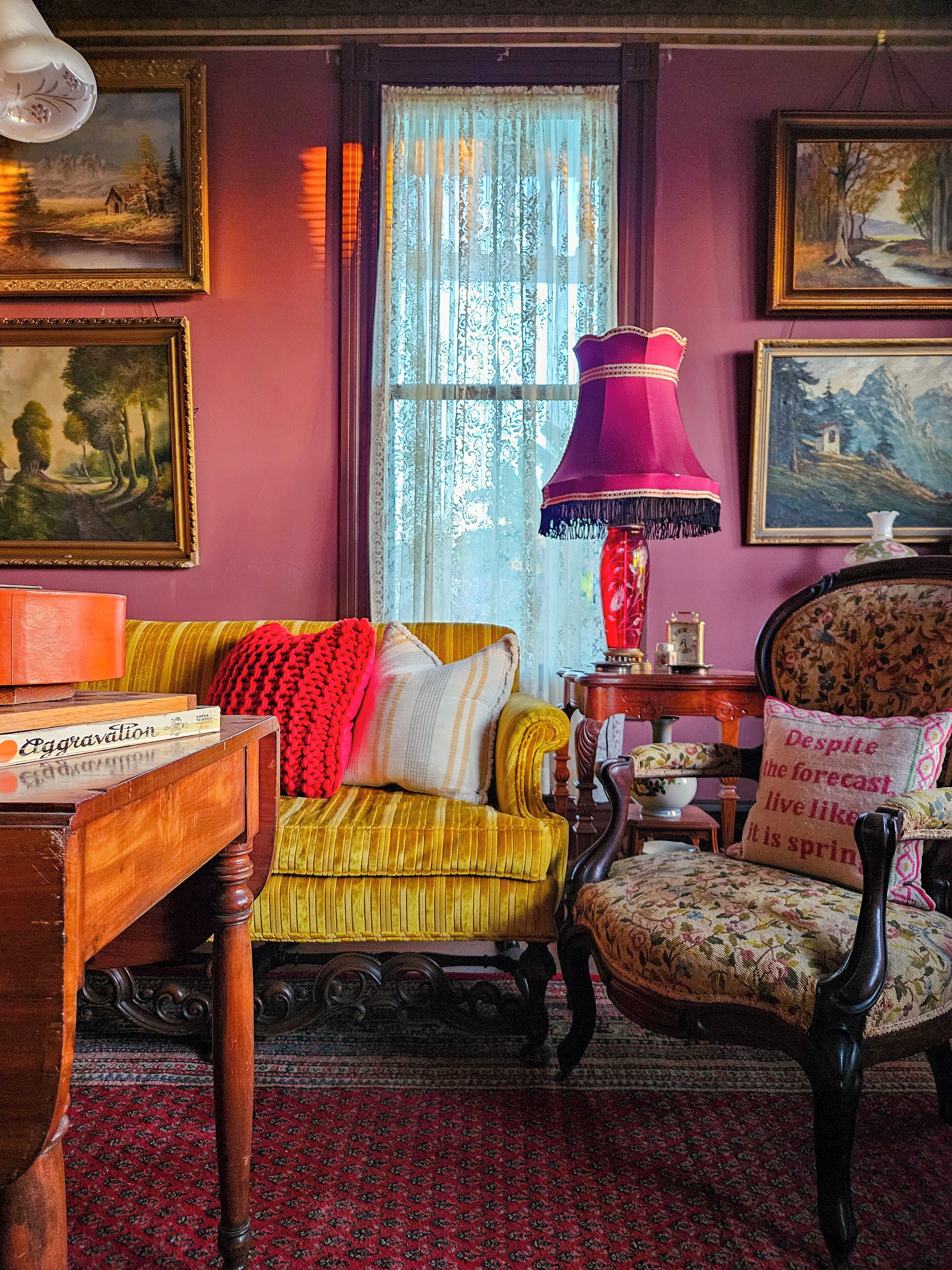 Une pièce comprenant un canapé, un fauteuil et une table avec des livres. Les murs sont ornés de tableaux et il y a une fenêtre avec des rideaux.